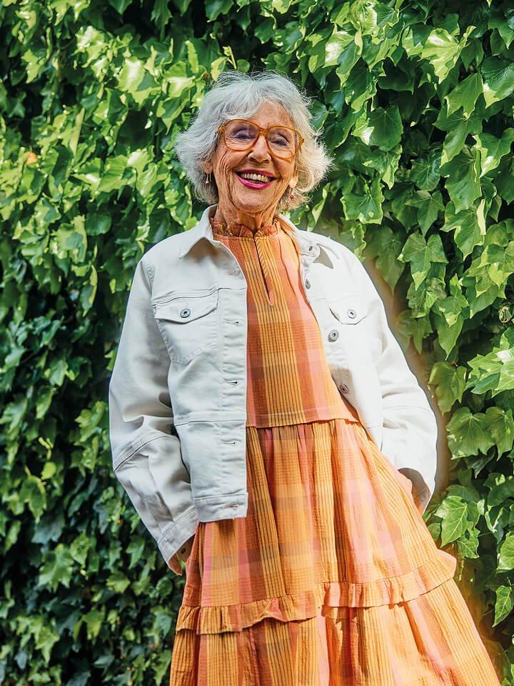 a woman in a dress and jacket standing next to a bush